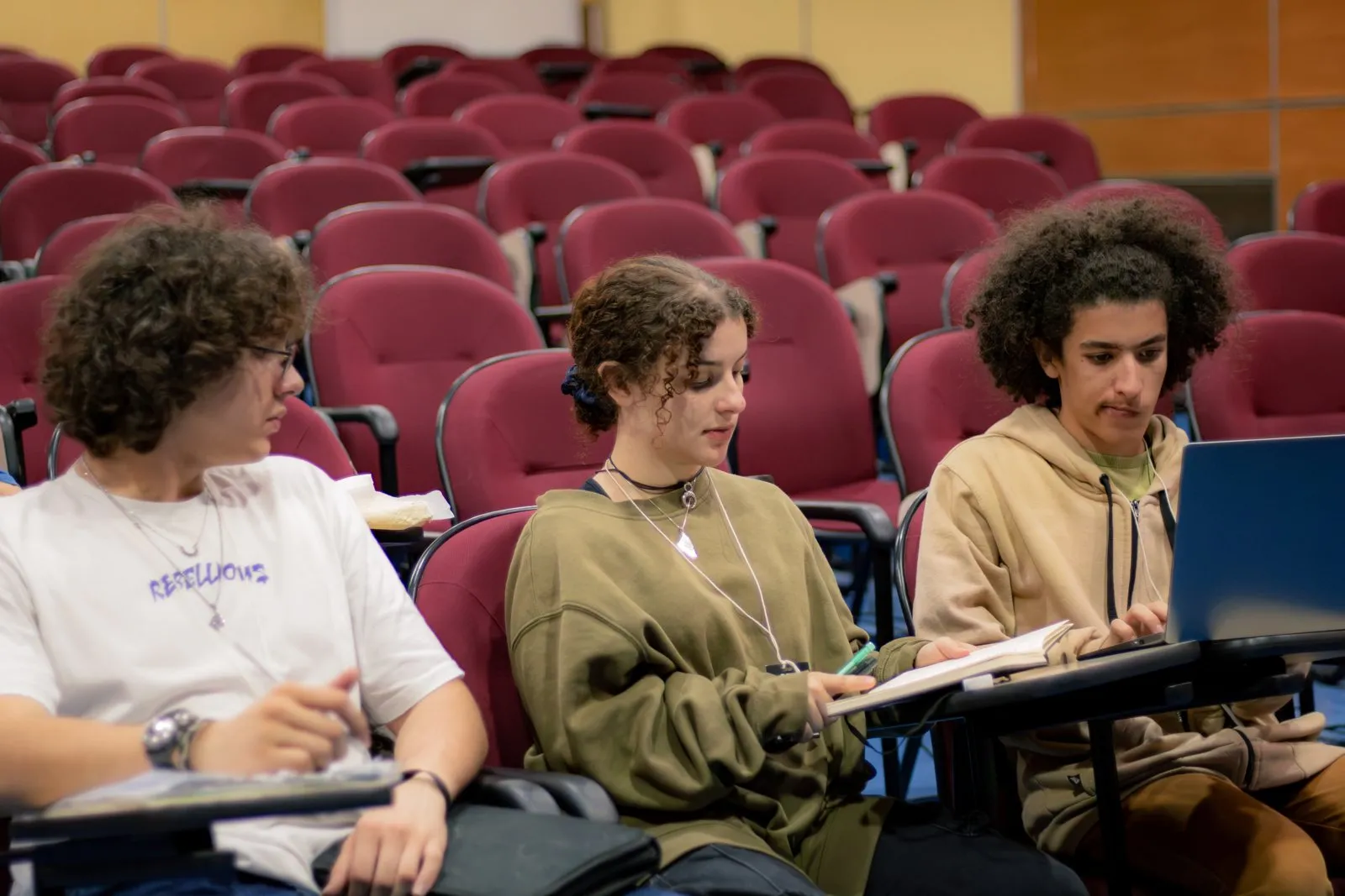 Três participantes trabalham com caderno e notebook no auditório da USP Leste.